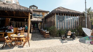 Un área de comedor al aire libre con mesas y sillas de techo de paja, un letrero que indica zonas 'STAY' y 'CO-WORK', y una tabla de surf apoyada en una pared.