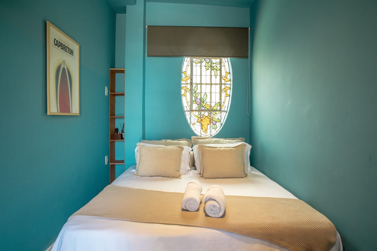 A hotel room with a bed, pillows, a window with a stained glass design, and a framed picture on the wall.