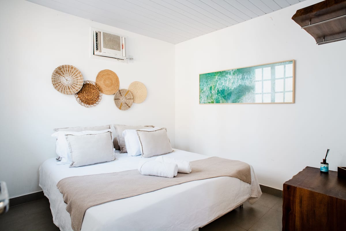 A neatly made bed with white linens and a beige throw blanket, a wooden nightstand with a lamp, and a wall-mounted air conditioner.