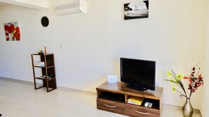 A living room with a flat-screen TV on a wooden stand, a small shelf with books, a clock, and a vase with flowers. - Stunning 2-bed Apartment in Poli Crysochous (Polis)
