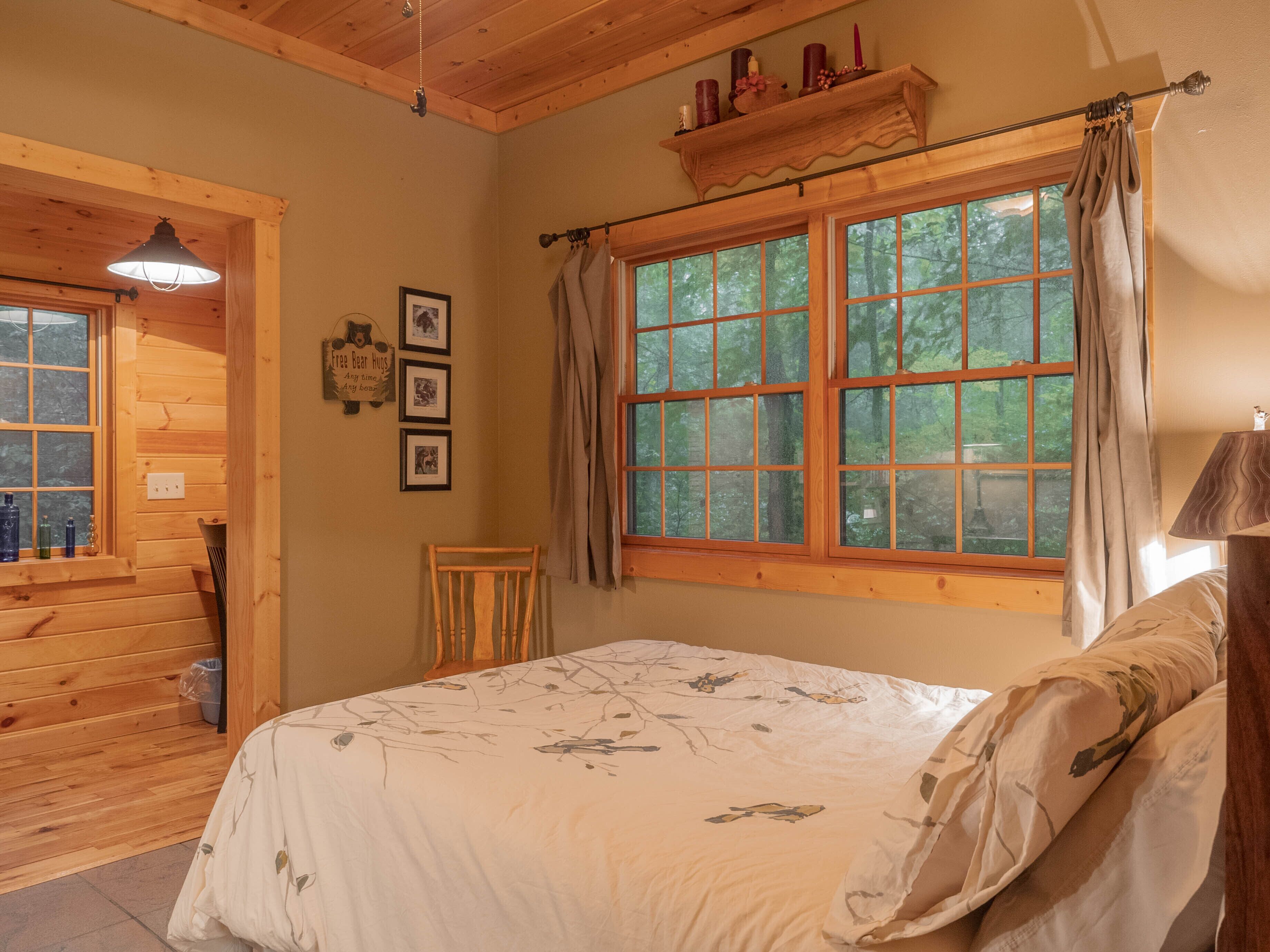 Un dormitorio con un ventanal grande, paredes de madera, una cama con ropa de cama floral, una silla y una estantería con velas.