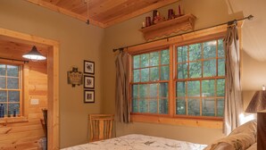 A bedroom with a large window, wooden walls, a bed with floral bedding, a chair, and a shelf with candles. - Smokies Hideaway -  2 bdrms/2 bath, private, SECLUDED, BONFIRE pit, Large driveway (Townsend)