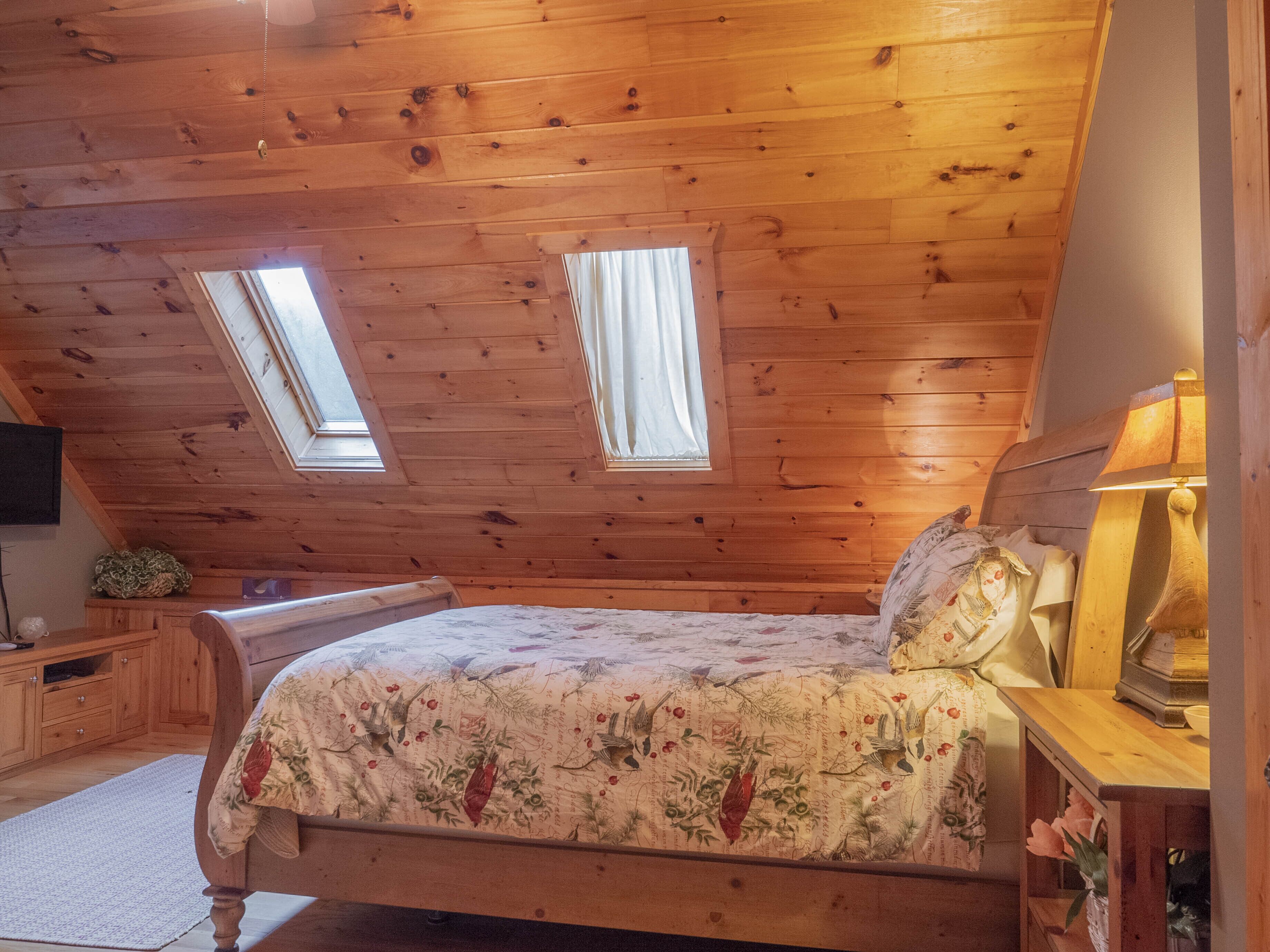 Ein gemütliches Schlafzimmer im Dachgeschoss mit Holzbalken, einem Bett mit Blumenbettwäsche, einem Nachttisch mit Lampe und zwei Dachfenstern.