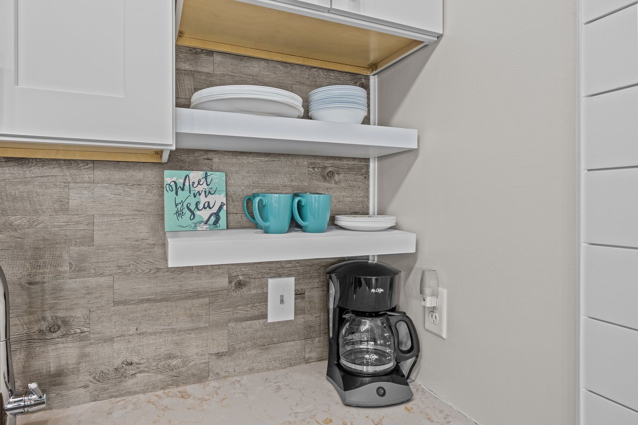 A kitchen with white cabinets, a wooden backsplash, a coffee maker, and teal cups on a shelf.