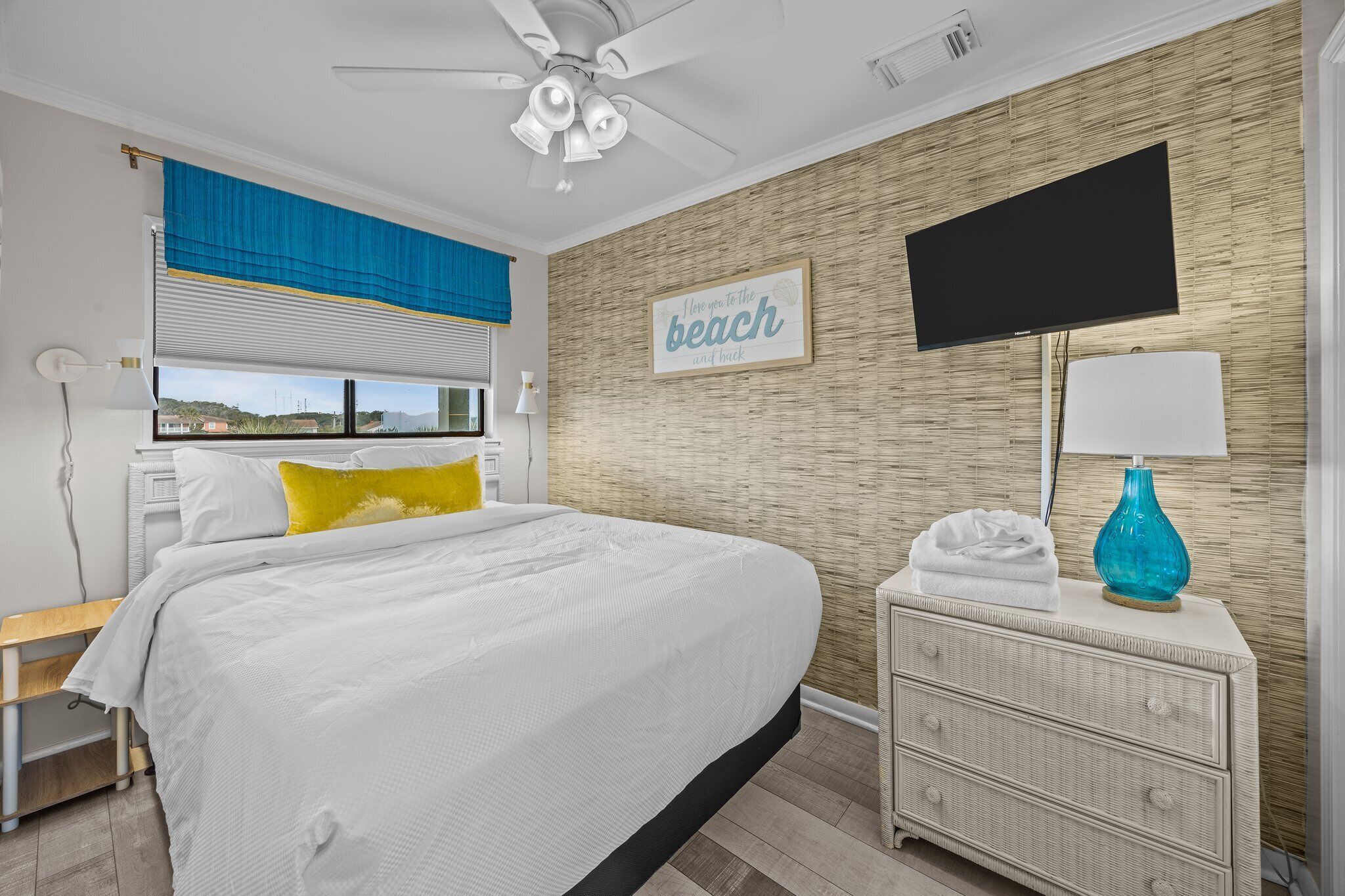 A bedroom with a bed, a ceiling fan, a TV, a bedside table, and a wall with a beach-themed sign.