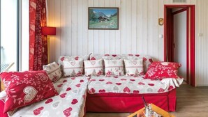 A cozy living room with a sectional sofa adorned with heart patterns, a framed landscape painting above it, and a red door to the right. - Baikonour by Interhome (Le Corbier)
