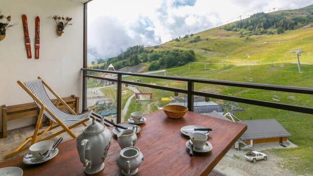 A balcony with a view of a green valley, a wooden chair, and a table with tea set. - Baikonour by Interhome (Le Corbier)