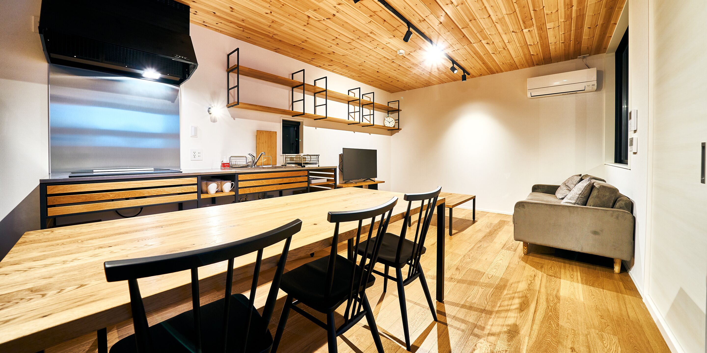 A modern kitchen with wooden flooring, a dining table with chairs, a sofa, and built-in shelves.