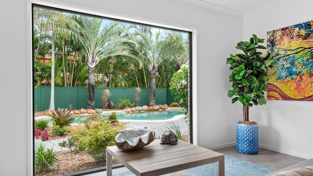 Una sala de estar moderna con un amplio ventanal corredera que da a una zona de piscina, una mesa de centro de madera, una alfombra azul y una planta en maceta.