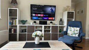A living room with a TV, bookshelves, a blue chair, and a vase with flowers on a table. - Hidden Gem - Bed & Breakfast (Upper Hutt)