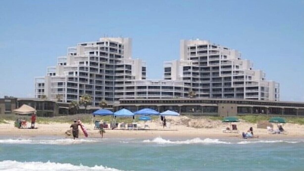 Un hotel grande con varios pisos y balcones, situado cerca de una playa con sombrillas y gente.