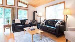 A cozy living room with two black leather sofas, a wooden coffee table, and a white armchair. Large windows provide natural light and a view of greenery outside. - Summer Basecamp: Near Mt Baker, BBQ, AC, Fireplace (Glacier)