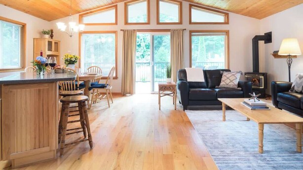 A spacious living area with a wooden ceiling, large windows, a fireplace, and a kitchen area with a bar. - Summer Basecamp: Near Mt Baker, BBQ, AC, Fireplace (Glacier)
