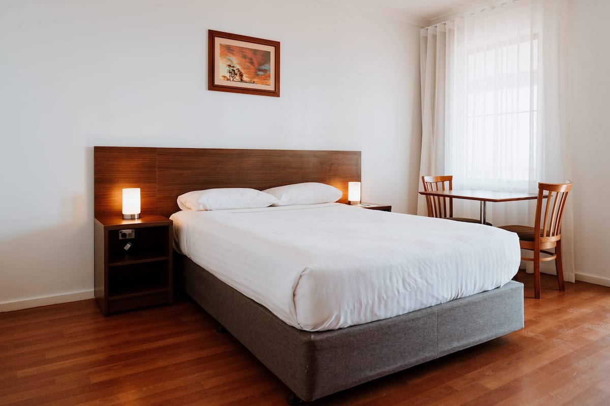 Een net opgemaakt bed met witte lakens, een houten hoofdbord en twee bedlampjes. Aan de rechterkant van het bed staat een kleine tafel met stoelen, en erboven hangt een schilderij aan de muur.