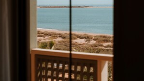 Vista desde una ventana a una playa con cielo despejado, orilla arenosa y mar tranquilo.
