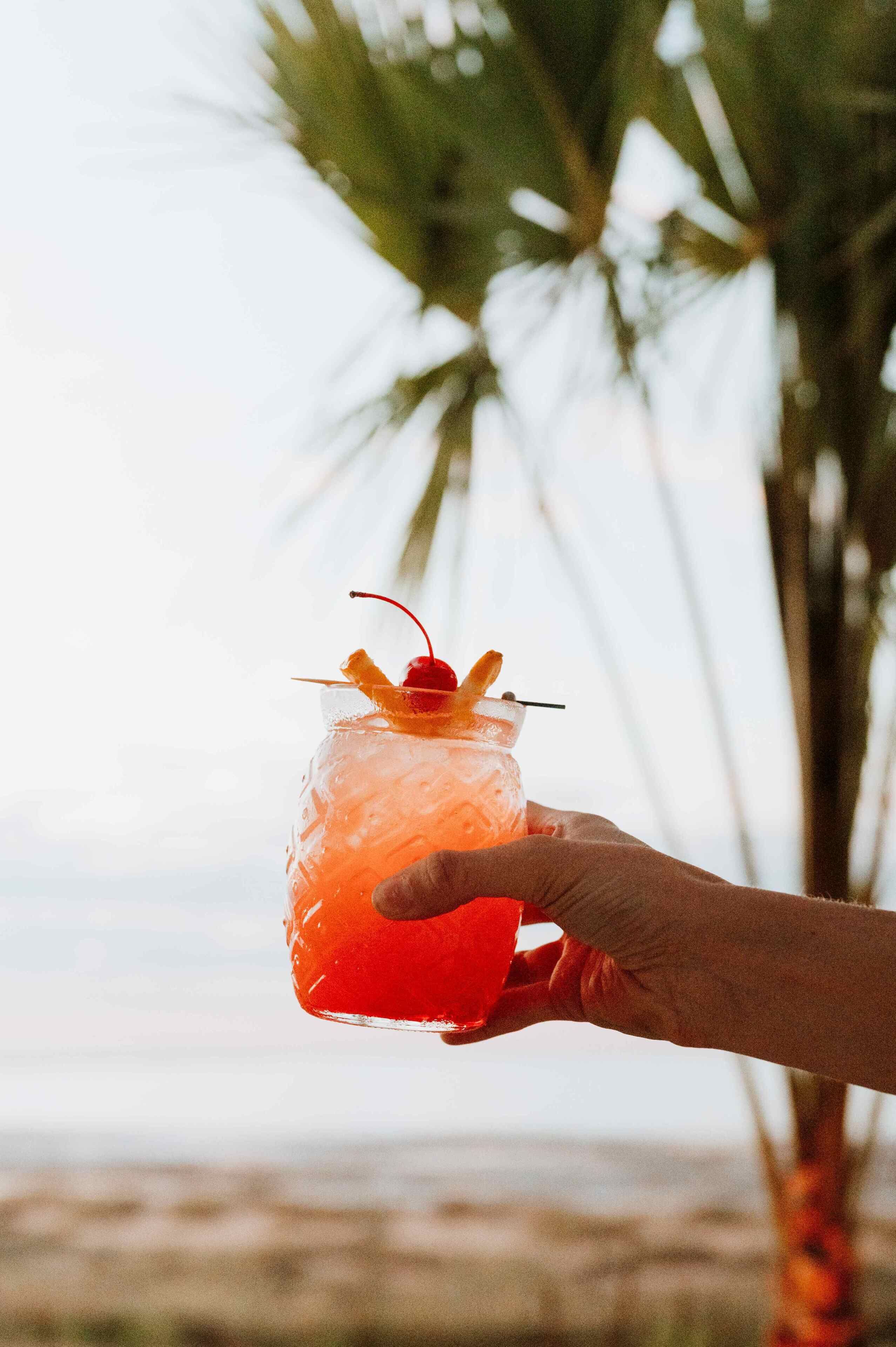 A hand holding a cocktail with a cherry garnish, palm trees in the background, and a blurred view of the ocean.