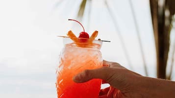 A hand holding a cocktail with a cherry garnish, palm trees in the background, and a blurred view of the ocean.