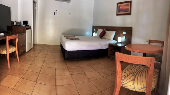 A hotel room with a bed, a desk, a chair, a small table, and a wall-mounted air conditioner.