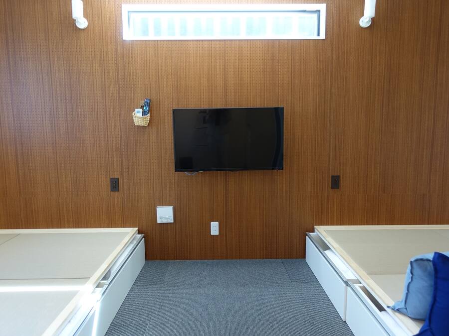 A room with a wooden paneled wall, a mounted flat-screen TV, a small basket, and a bed with a blue blanket.