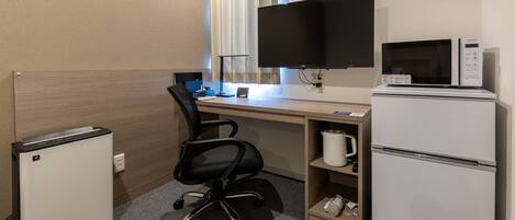 A hotel room with a desk, a black office chair, a flat-screen TV, a microwave, a mini-fridge, and a coffee maker.