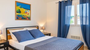 A bedroom with a bed, a nightstand, a lamp, a window with curtains, and a painting on the wall.