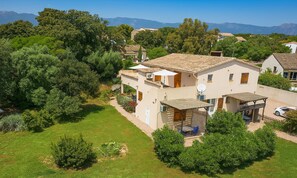 A two-story house with a tiled roof, surrounded by greenery and mountains in the background.