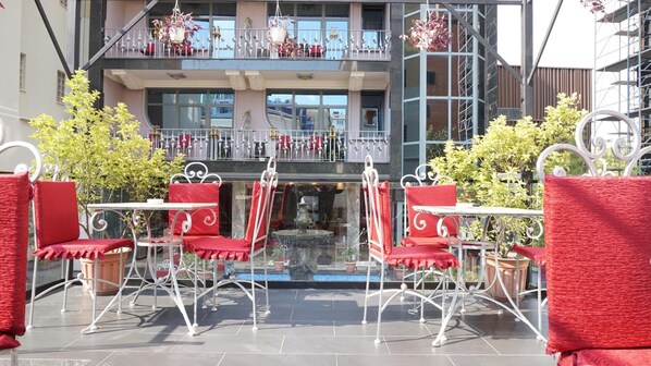 Una terraza con sillas rojas y una mesa, una fuente decorativa y vistas a un edificio con balcones.