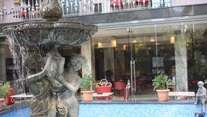 Une fontaine ornée d’une statue représentant une femme et un enfant, entourée d’un bassin. On y trouve également des plantes en pot et des sièges d’extérieur.