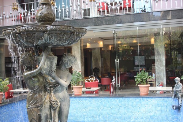 A fountain with a statue of a woman and a child, surrounded by a pool. There are potted plants and outdoor seating.