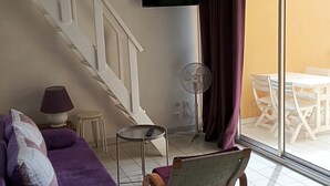 A living room with a purple sofa, a small table with a lamp, a fan, and a TV mounted on the wall.