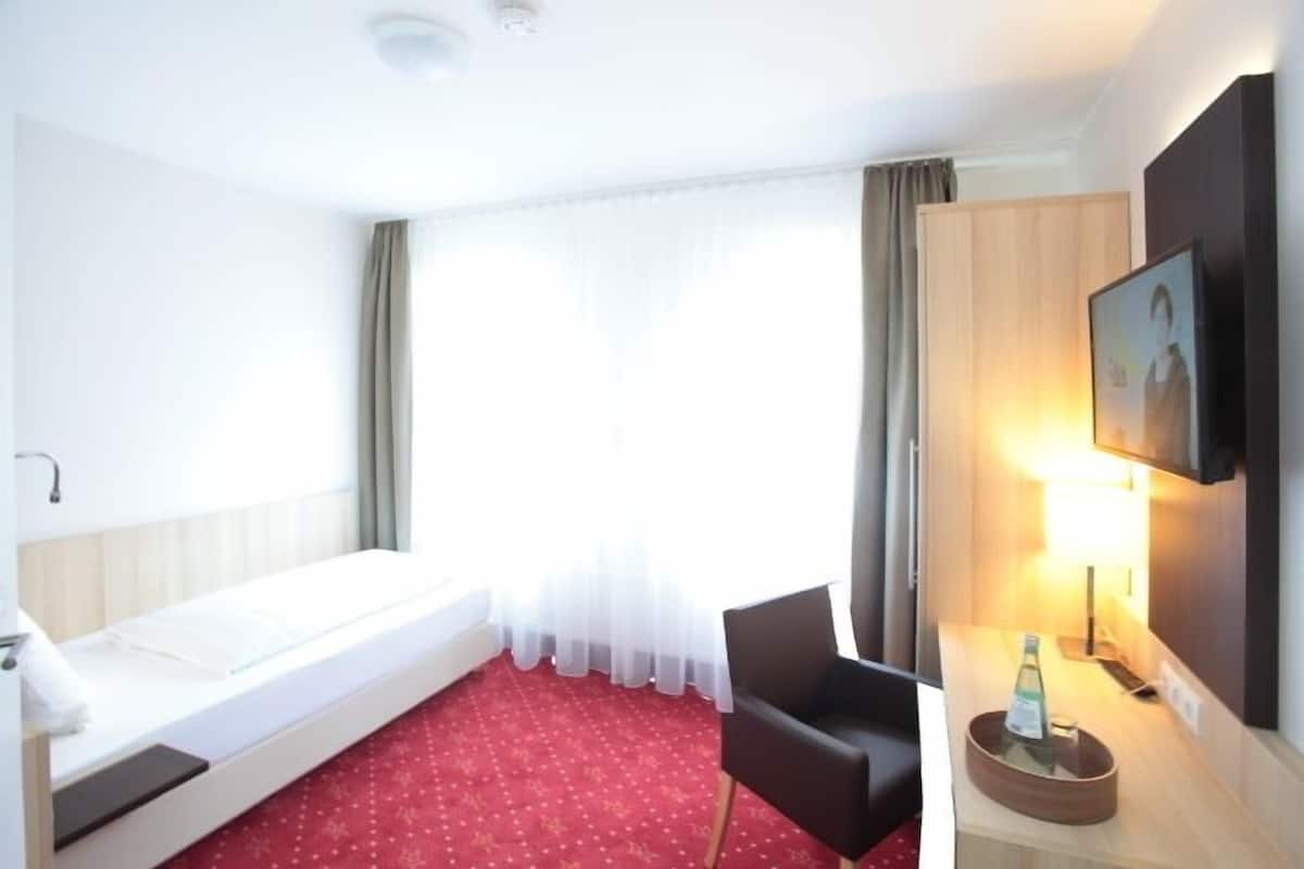 Habitació d'hotel amb un llit individual, una taula de treball amb una làmpada, una cadira i un televisor penjat a la paret.