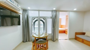 A room with a round glass table, a TV mounted on the wall, a sliding glass door leading to a bedroom, and a wooden bench. - New Hampton Hospitality - Cornelia Hotel & Apartment (Ho Chi Minh City)