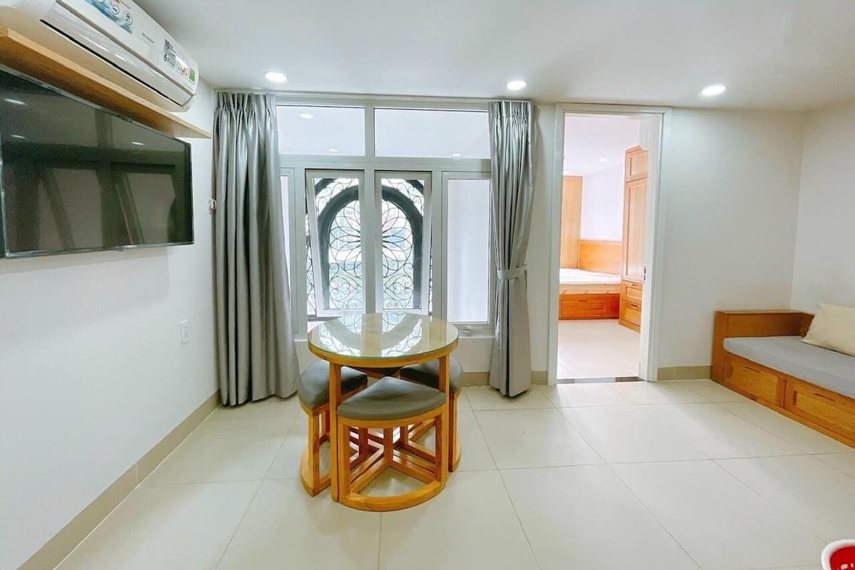 A room with a round glass table, a TV mounted on the wall, a sliding glass door leading to a bedroom, and a wooden bench.