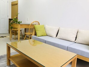 A modern living room with a glass-top coffee table, a grey sofa with white cushions, a wooden dining table with chairs, and a wooden door.
