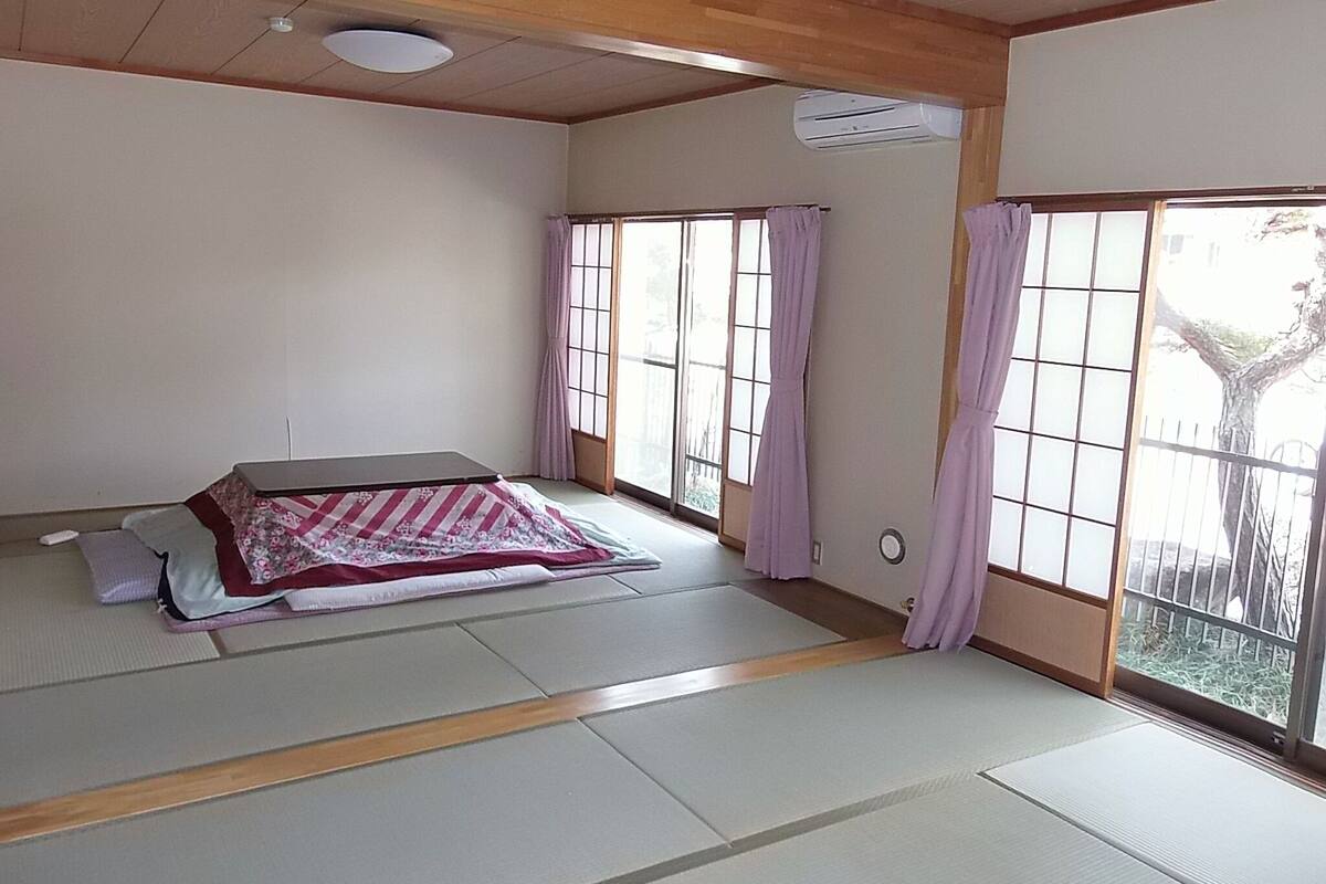 A traditional Japanese room with tatami flooring, a futon bed, sliding shoji doors, and a view of a tree outside.