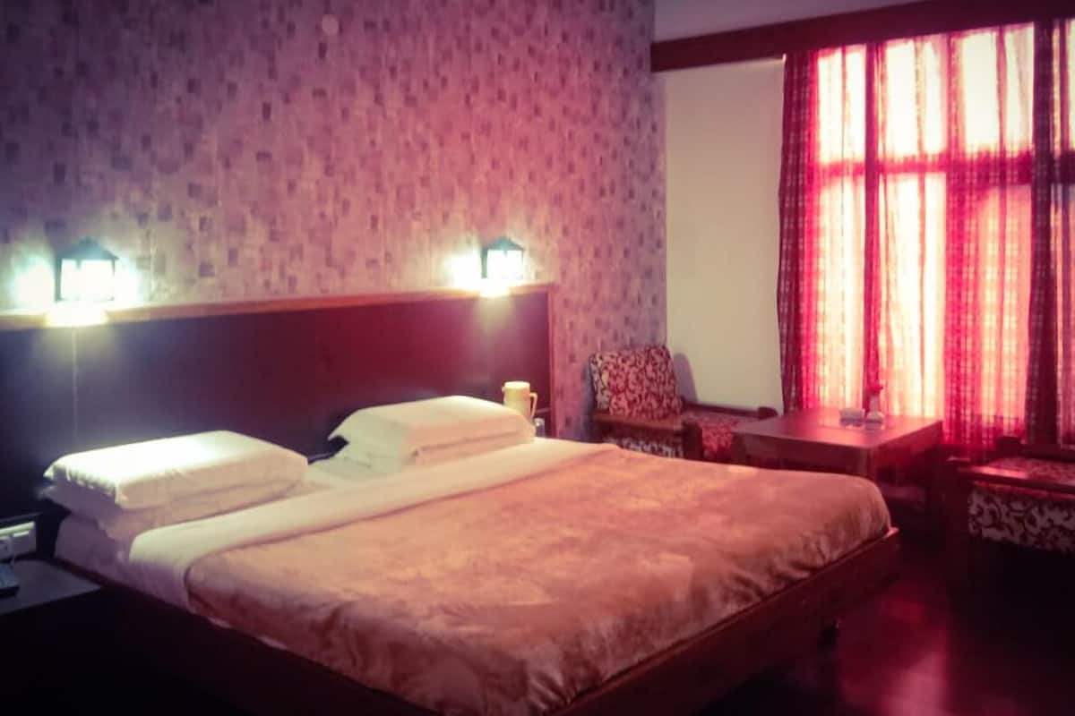 Quarto de hotel com uma cama grande, duas luminárias de cabeceira, uma mesa de apoio, uma cadeira e uma janela com cortinas vermelhas.