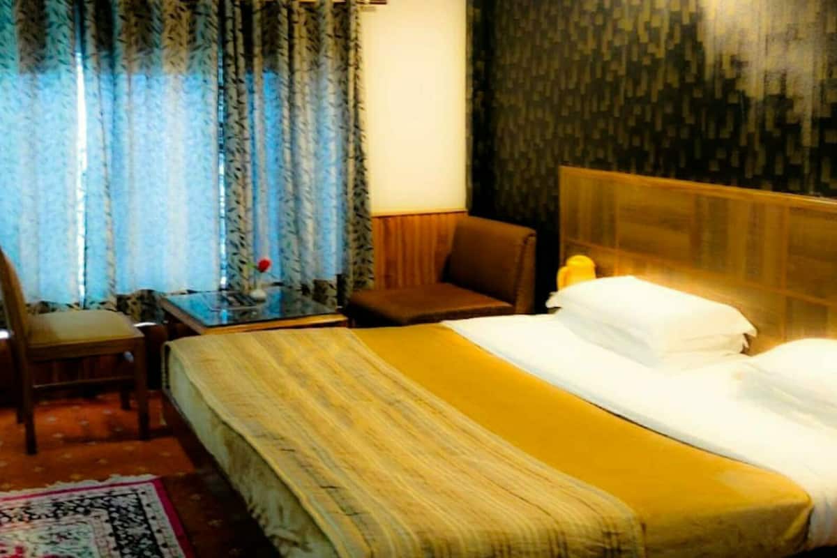 Quarto de hotel com cama, uma cadeira, uma mesa pequena e cortinas.