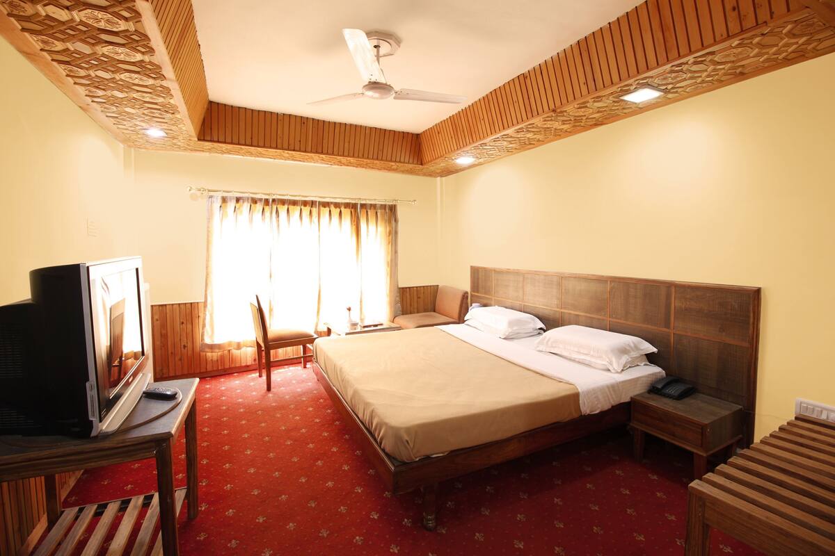 Quarto de hotel com cama, televisão, escrivaninha, cadeira, mesa de cabeceira e ventoinha no teto.