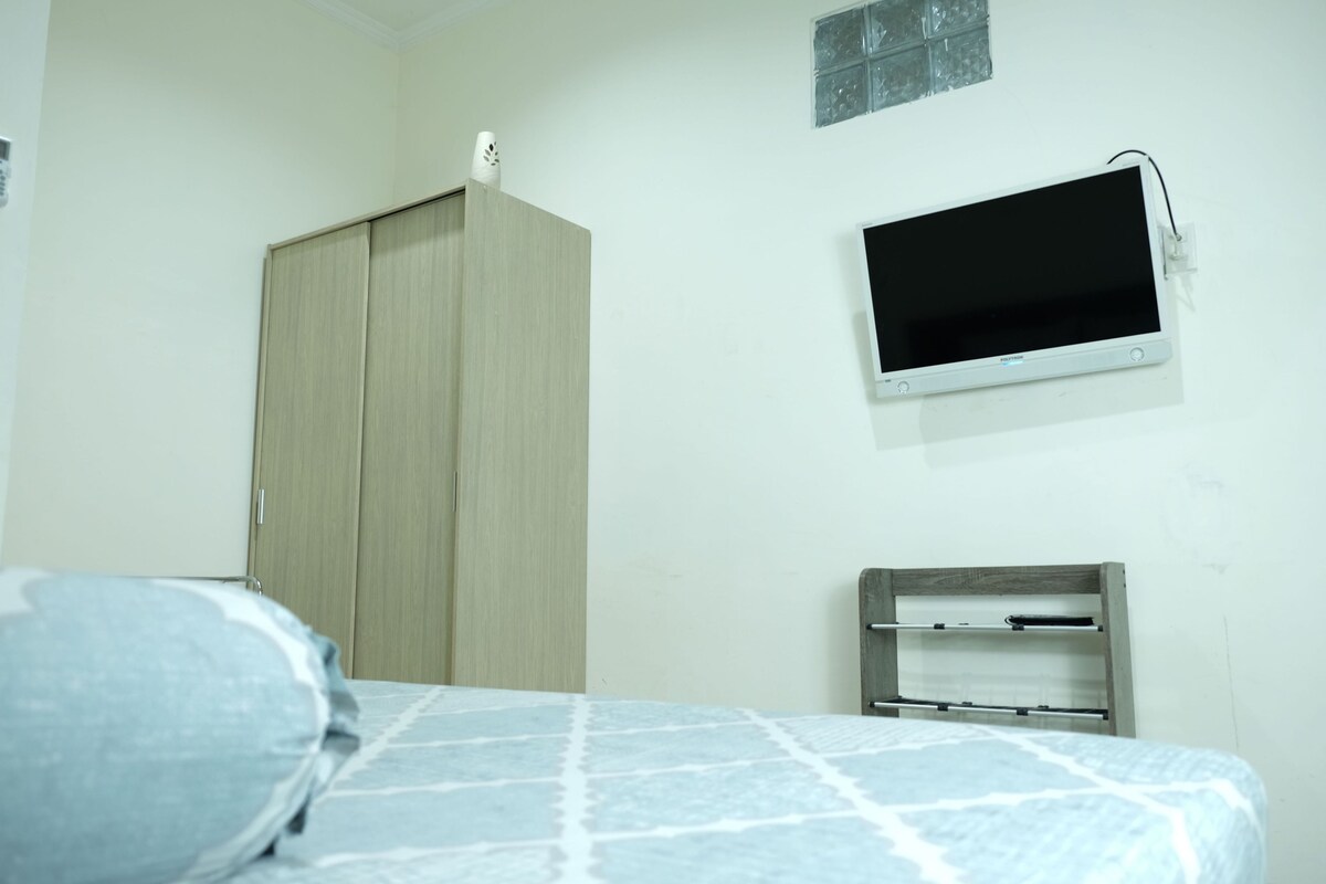 Un dormitorio con cama, armario, televisor montado en la pared y una pequeña estantería.