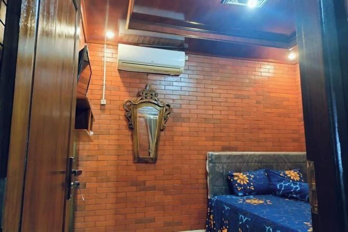 A room with a brick wall, a bed with patterned bedding, a mirror, and an air conditioning unit.