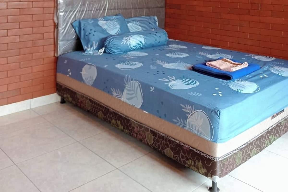 A room with a brick wall, a bed with a blue mattress and patterned bedding, a single pillow, and a small table with a magazine.