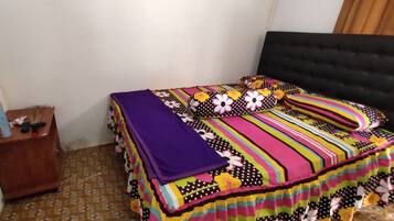 A bedroom with a bed covered in a colorful striped blanket, a bedside table with a lamp, and a standing fan.