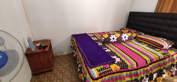 A bedroom with a bed covered in a colorful striped blanket, a bedside table with a lamp, and a standing fan. - Limasan Mbah Darmo (Tepus)