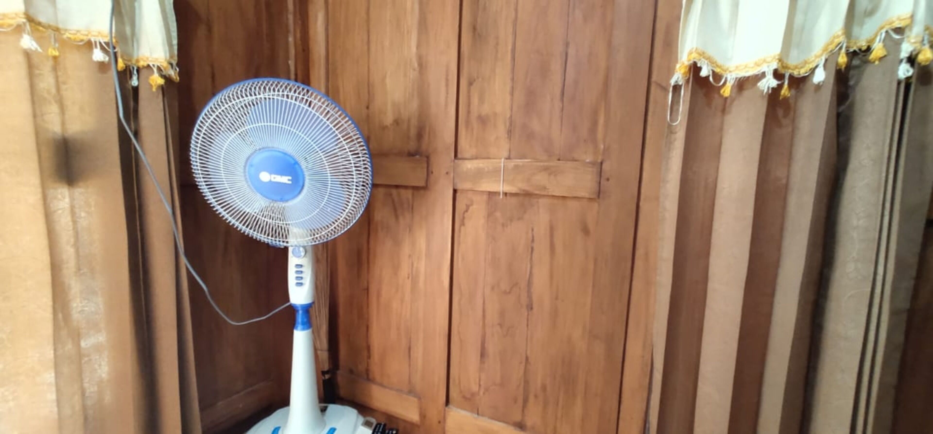 A standing fan in a corner with wooden walls and curtains.