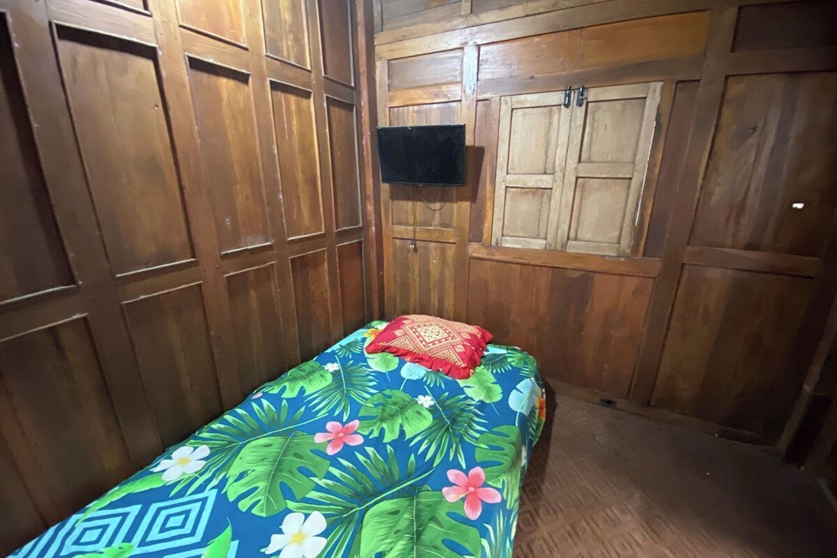 Una habitación pequeña con paneles de madera, una cama individual con colcha estampada y un televisor montado en la pared.