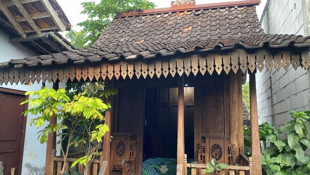 Une construction traditionnelle en bois, avec un toit en tuiles, une véranda en bois dotée de deux chaises et une porte donnant sur l’intérieur.