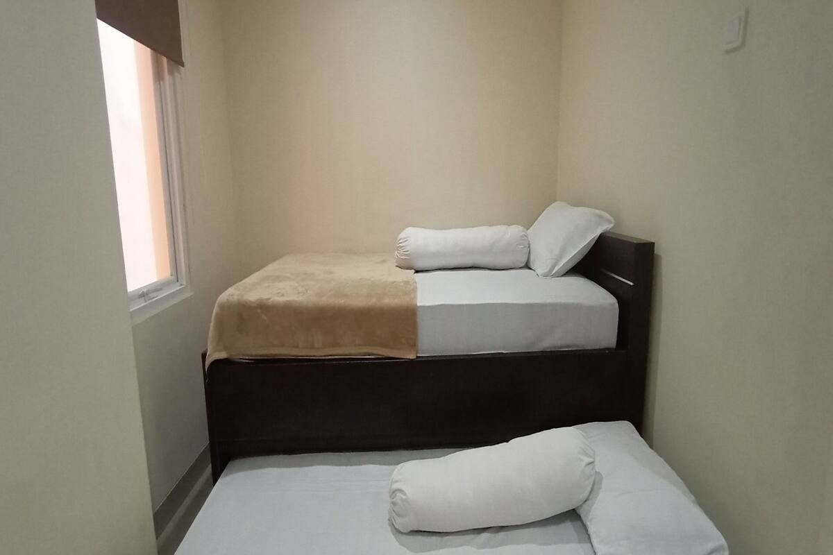 A small, single-bed room with a window on one side, a dark headboard, and a white bedspread.