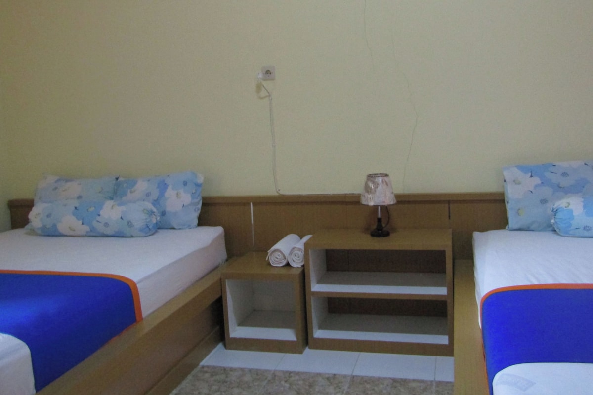 A small, simple room with two beds, each with a blue and white floral pillow, a small nightstand with a lamp, and a power outlet on the wall.