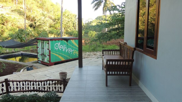 A covered outdoor area with a bench, a green container with 'deliver' written on it, and a view of a natural landscape with trees and a water feature.