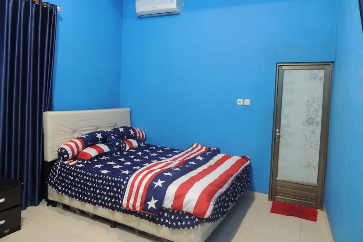 A bedroom with a bed covered in an American flag-themed blanket, a blue wall, an air conditioning unit, a wardrobe, and a red rug.